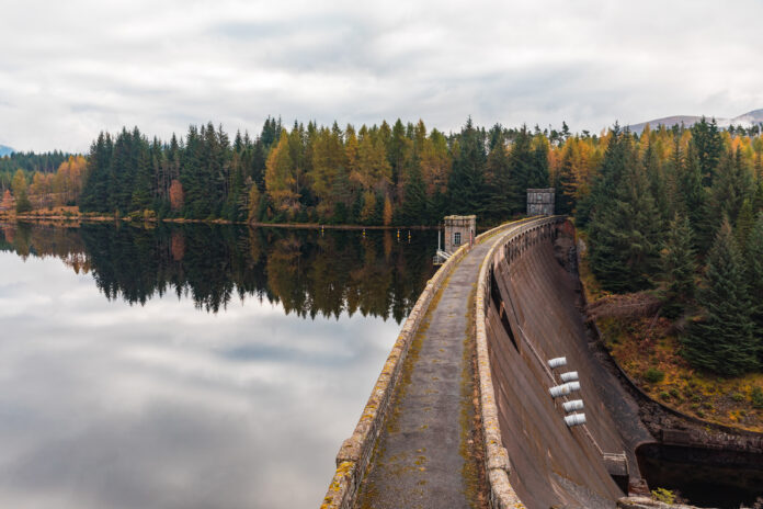 Dam wall curving around reservoir reflecting autumnal forest in scottish highlands
